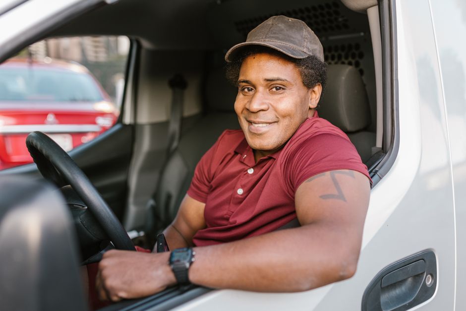 Smiling delivery driver wearing a red polo shirt and brown cap, sitting in vehicle. Professional service.